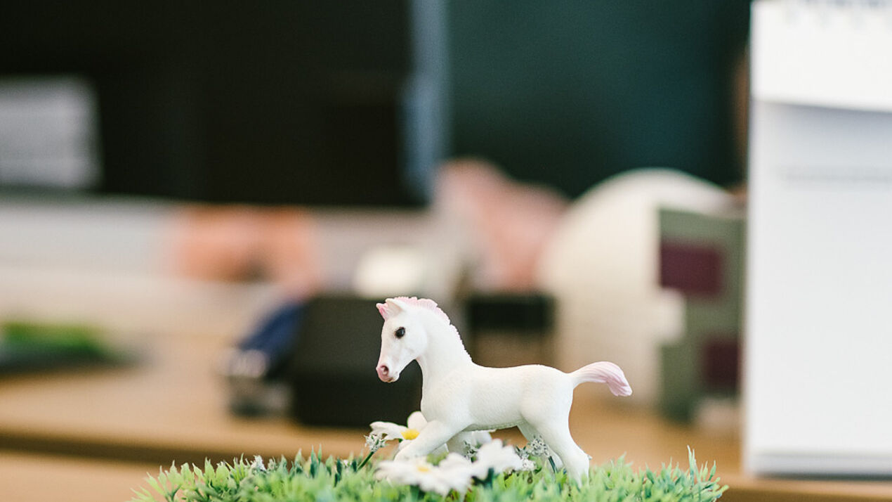 Kleine weiße Pferdefigur steht auf künstlichem Gras auf einem Schreibtisch, im Hintergrund sind unscharf Bürogegenstände zu erkennen.