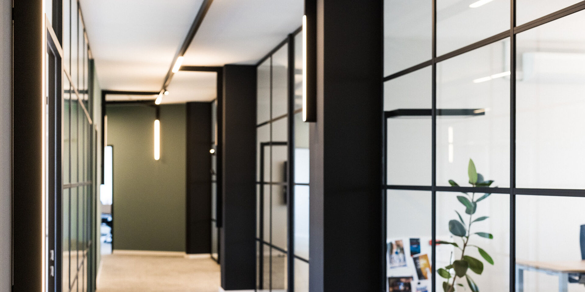 Moderner Büroflur mit Glaswänden, dunklen Rahmen und linearen LED-Leuchten, gestaltet in minimalistischem, zeitgemäßem Design.